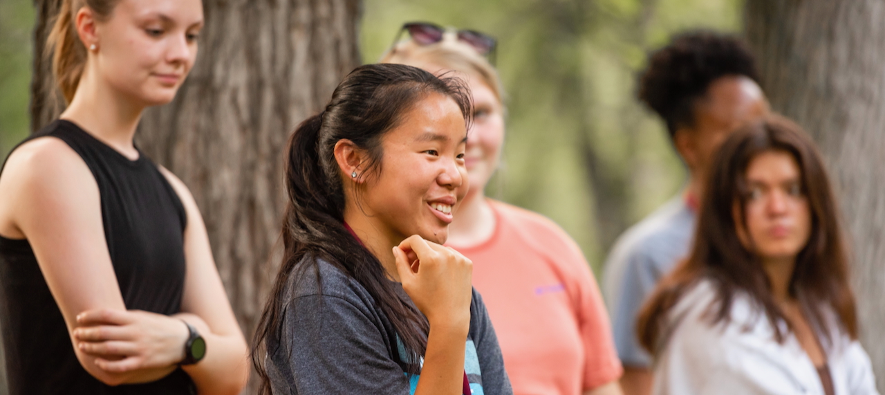 Smiling UMR student speaking outdoors, surrounded by a diverse group of people in casual clothing near trees