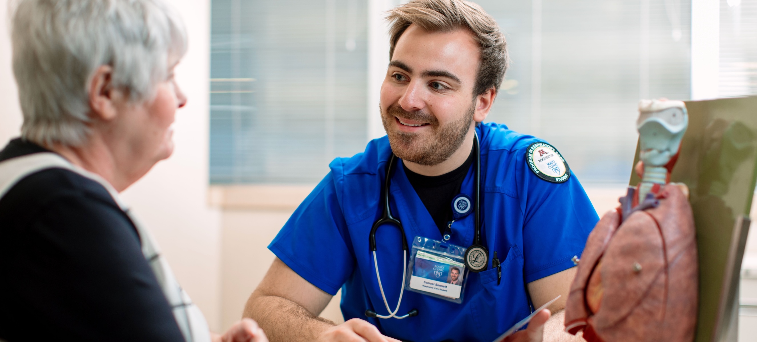 A young male nurse in blue scrubs smiles while speaking with an older woman during a medical consultation.