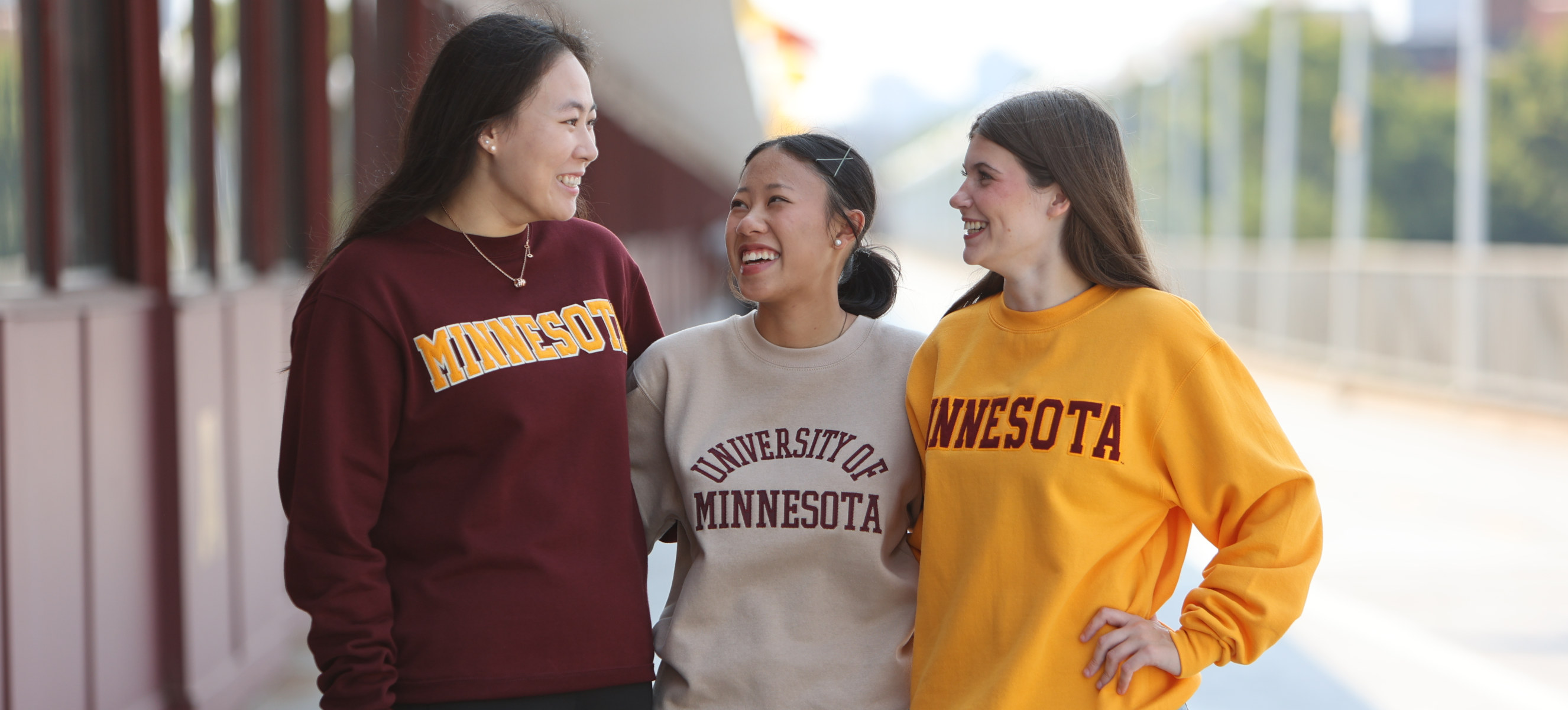 Three students in University of Minnesota sweatshirts smile and talk outside on a sunny day.