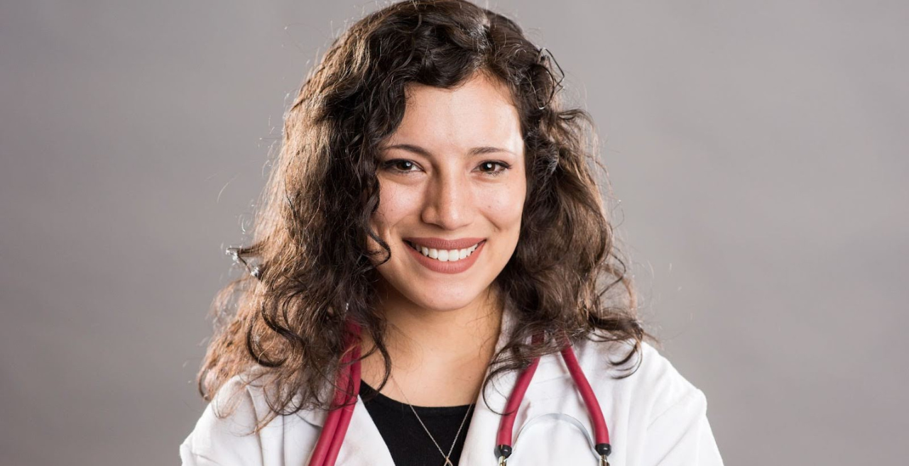 Smiling medical professional with curly hair wears a white lab coat and stethoscope against a neutral background.