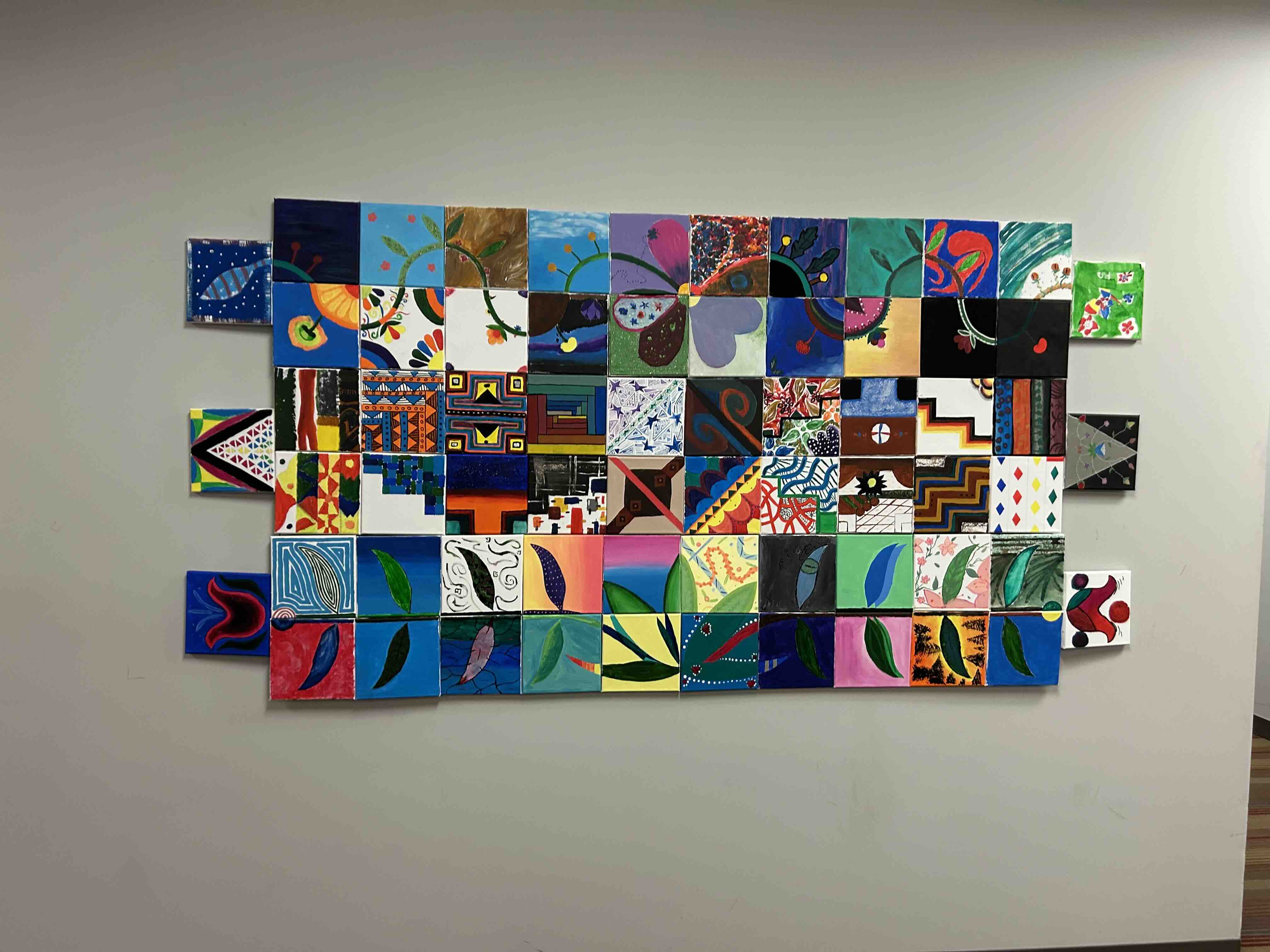 Large collaborative mural made of colorful square paintings arranged in a grid on a light wall, featuring abstract, geometric, and organic designs in vibrant hues.