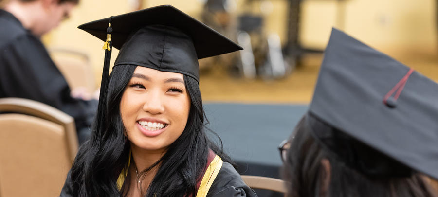 Female graduate sitting at a table with classmate and smiling with cap and gown on. 